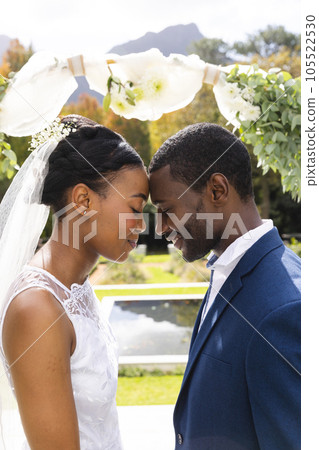 Happy african american bride and groom touching heads and smiling under wedding arch in sunny garden 105522530