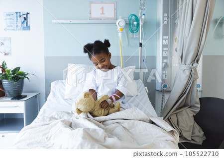 Happy african american girl patient lying on bed playing with teddy bear in patient room at hospital 105522710