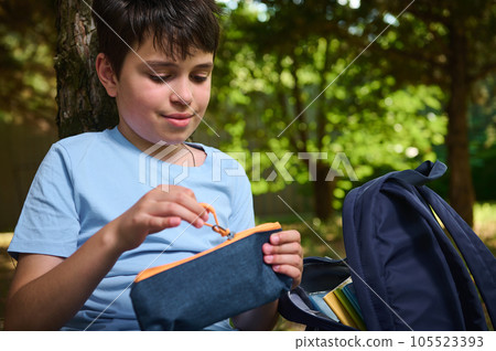 Caucasian teenage boy, smart primary school student opening his pencil case while doing homework in the park after class Caucasian teenage boy, smart primary school student opening his pencil case while doing homework in the park after class 105523393