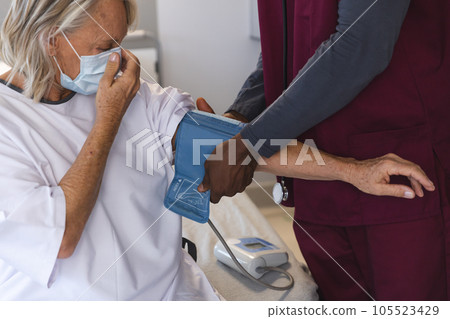 African american doctor measuring blood pressure of senior caucasian patient, wearing face masks 105523429
