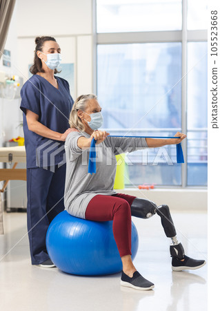 Caucasian physiotherapist and senior woman with artificial leg with face masks using exercise band 105523668