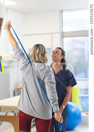 Caucasian female physiotherapist and senior woman using exercise band stretching arms 105523676
