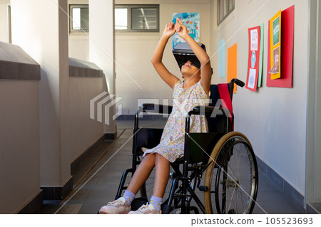 Happy biracial schoolgirl sitting in wheelchair and using vr headset at elementary school corridor 105523693