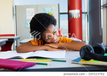 Happy biracial schoolboy sitting at desk and lying on books in classroom 105523762