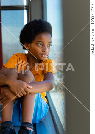 Thoughtful biracial schoolboy wearing yellow tshirt and looking through window in classroom 105523779