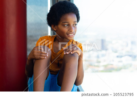 Happy biracial schoolboy wearing yellow tshirt and looking through window in classroom 105523795