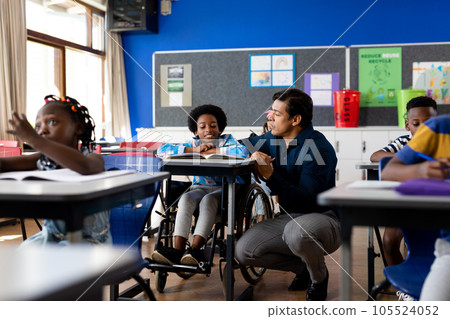 Biracial male teacher helping schoolboy sitting in wheelchair in classroom at elementary school 105524052