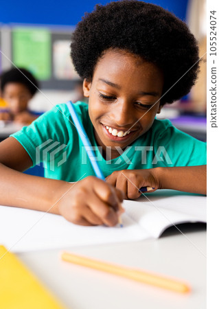 Portrait of happy african american elementary schoolboy writing in notebook at desk in class 105524074
