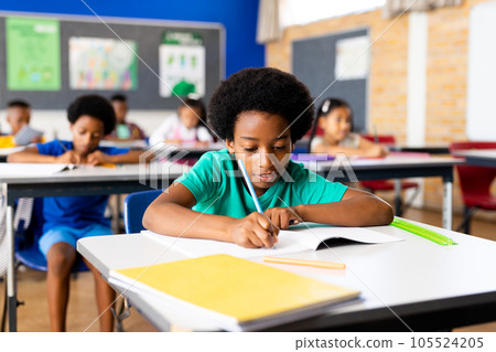 Portrait of happy african american elementary schoolboy writing in notebook at desk in class 105524205