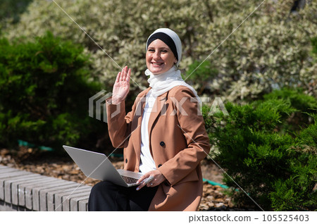 Portrait of young woman in hijab using laptop outdoors. Muslim woman looking at the camera and waving. Portrait of young woman in hijab using laptop outdoors. Muslim woman looking at the camera and waving. 105525403