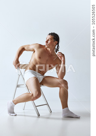 Handsome, stylish mature man posing shirtless in underwear, sitting on chair against grey studio background. Muscular body 105526505