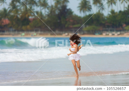 A slender girl in white clothes runs along the beach against the background of the sea. 105526657