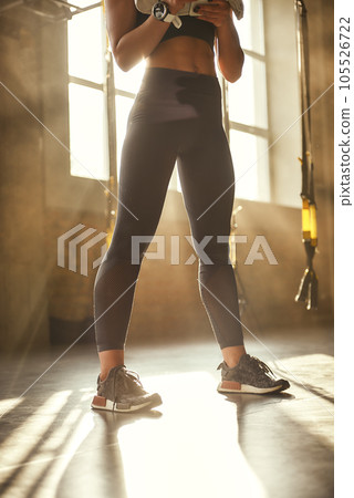 Great shape. Close-up of young athletic woman with perfect body in sport leggings is standing in front of window in professional semi-dark gym. 105526722