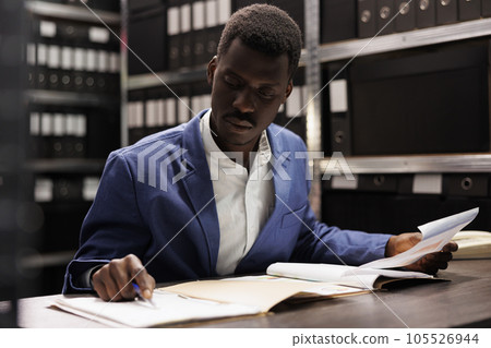 African american officer analyzing police report, searching for new clues trying to catch mysterious suspect. Private detective working overhours at crime case, checking evidene in arhive room 105526944