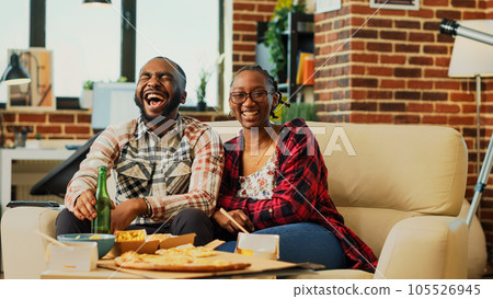 Smiling people laughing at comedy film on television, feeling happt spending time together. Young life partners enjoying movie on tv, eating chips and takeaway food. Tripod shot. 105526945