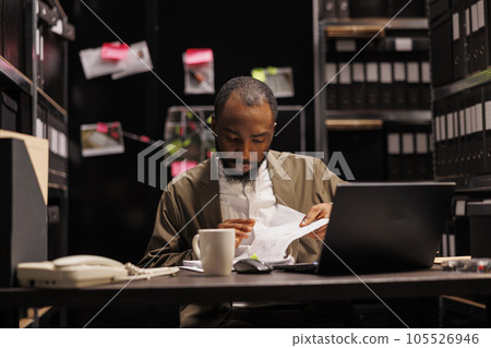 African american police detective searching evidence in crime case file. Private investigator reading witness statement in reports while working overtime and sitting at desk at night time 105526946