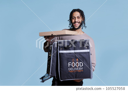 Happy arab courier holding pizza box and looking at camera with cheerful facial expression. Carefree pizzeria delivery service worker taking fast food from thermal backpack portrait 105527058