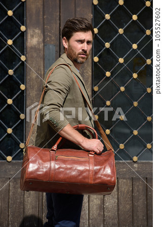 a man with a bag. black leather travel bag, street photo. man is wearing blue sneakers, jeans, a white T-shirt and a jacket a man with a bag. black leather travel bag, street photo. man is wearing blue sneakers, jeans, a white T-shirt and a jacket 105527602