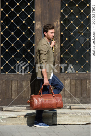 a man with a bag. black leather travel bag, street photo. man is wearing blue sneakers, jeans, a white T-shirt and a jacket a man with a bag. black leather travel bag, street photo. man is wearing blue sneakers, jeans, a white T-shirt and a jacket 105527603