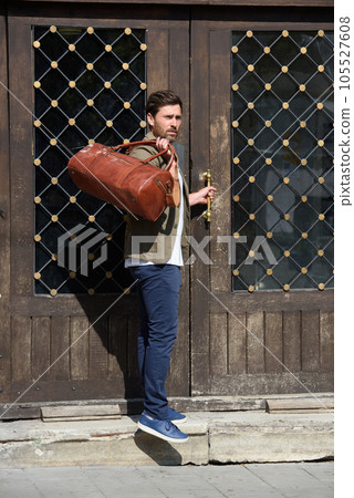 a man with a bag. black leather travel bag, street photo. man is wearing blue sneakers, jeans, a white T-shirt and a jacket 105527608