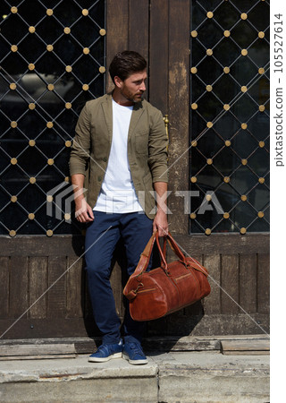 a man with a bag. black leather travel bag, street photo. man is wearing blue sneakers, jeans, a white T-shirt and a jacket a man with a bag. black leather travel bag, street photo. man is wearing blue sneakers, jeans, a white T-shirt and a jacket 105527614