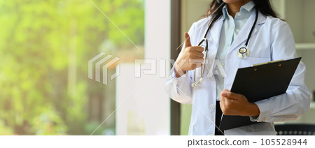 Doctor dressed in white overall with stethoscope holding clipboard and showing thumbs up Doctor dressed in white overall with stethoscope holding clipboard and showing thumbs up 105528944
