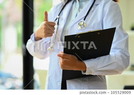 Doctor dressed in white overall with stethoscope holding clipboard and showing thumbs up 105529009