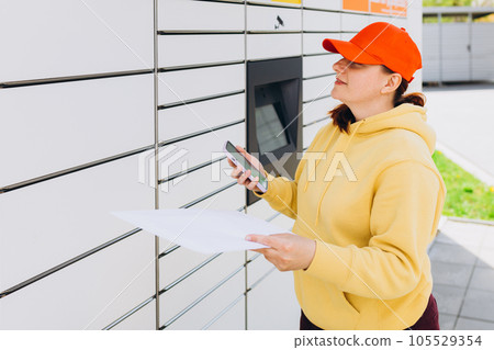 Young woman in red hat picks up parcel from automatic post office machine, Courier standing with phone and small box. Concept of fast delivery to automatic self lockers 105529354