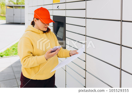 Young woman in red hat picks up parcel from automatic post office machine, Courier standing with phone and small box. Concept of fast delivery to automatic self lockers 105529355