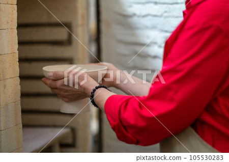 Craftswoman placing clay mugs into kiln for firing pottery while working in workshop. Craftswoman placing clay mugs into kiln for firing pottery while working in workshop. 105530283