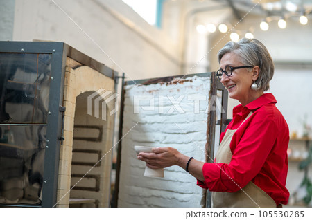 Positive craftswoman standing in front of open pottery kiln working in pottery workshop. Positive craftswoman standing in front of open pottery kiln working in pottery workshop. 105530285