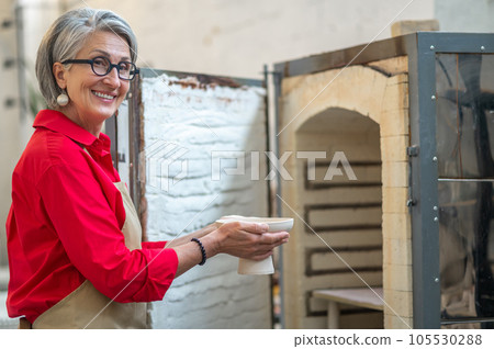 Craftswoman placing clay mugs into kiln for firing pottery while working in workshop. 105530288