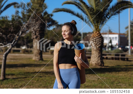 Young sporty woman preparing for training, yoga practice, pilates outdoors. Girl in headphones and sportswear with mat working out on summer vacation at park with palm trees. Sports, healthy lifestyle Young sporty woman preparing for training, yoga practice, pilates outdoors. Girl in headphones and sportswear with mat working out on summer vacation at park with palm trees. Sports, healthy lifestyle 105532458