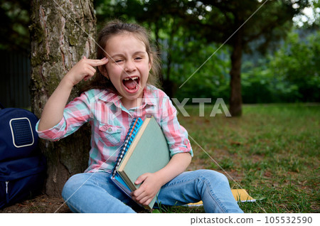 Mischievous little child girl sitting under a tree, pointing her finger at her temple, making faces looking at camera Mischievous little child girl sitting under a tree, pointing her finger at her temple, making faces looking at camera 105532590
