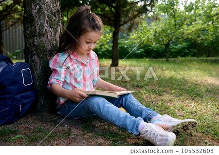 Little school girl reads a book in the park, sitting under tree with her backpack. Kids. Erudition. Education. Lifestyle Little school girl reads a book in the park, sitting under tree with her backpack. Kids. Erudition. Education. Lifestyle 105532603