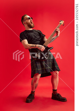 Bearded man in sunglasses and skirt, kilt playing guitar against red studio background. Rock and roll Bearded man in sunglasses and skirt, kilt playing guitar against red studio background. Rock and roll 105532781
