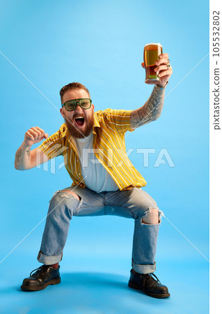 Portrait of emotional and excited bearded man in sunglasses and yellow shirt posing with beer against blue studio background Portrait of emotional and excited bearded man in sunglasses and yellow shirt posing with beer against blue studio background 105532802
