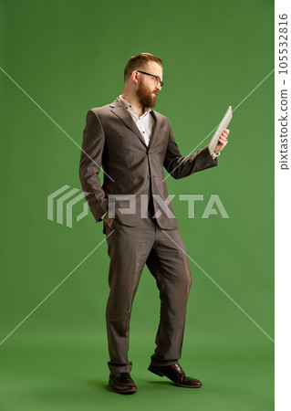 Full-length portrait of bearded businessman in suit and glasses seriously looking on tablet against green studio background Full-length portrait of bearded businessman in suit and glasses seriously looking on tablet against green studio background 105532816