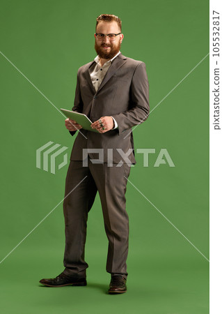 Full-length portrait of bearded stylish businessman in suit and glasses posing with tablet, looking at camera against green studio background Full-length portrait of bearded stylish businessman in suit and glasses posing with tablet, looking at camera against green studio background 105532817
