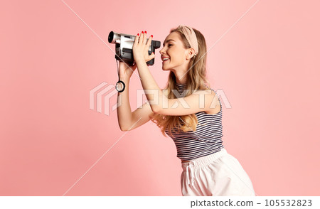 Woman dressed stylishly looking at handheld vintage camera and filming something against light pink background. Side view. 105532823