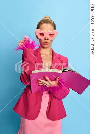 Portrait of charming girl, woman wearing glamor in unusual glasses, holding textbook looking at camera on light blue studio background. Fashion. 105532910
