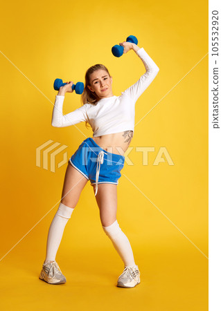 Full lenght of blonde girl trying to raise dumbbell looking at camera over bright yellow background. 105532920