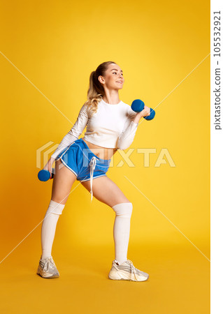 Sporty woman in sportswear holding dumbbells training muscles of hand and legs looking away over bright yellow background. Sporty woman in sportswear holding dumbbells training muscles of hand and legs looking away over bright yellow background. 105532921