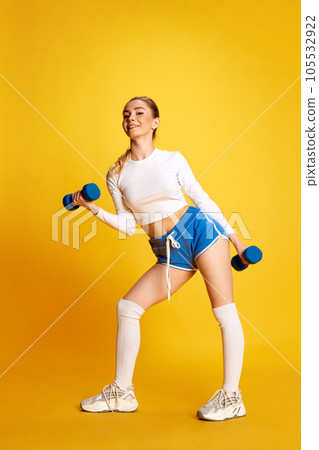 Full lenght image of young woman in training pumping up muscles of hands and legs looking at camera over yellow background. Side view Full lenght image of young woman in training pumping up muscles of hands and legs looking at camera over yellow background. Side view 105532922