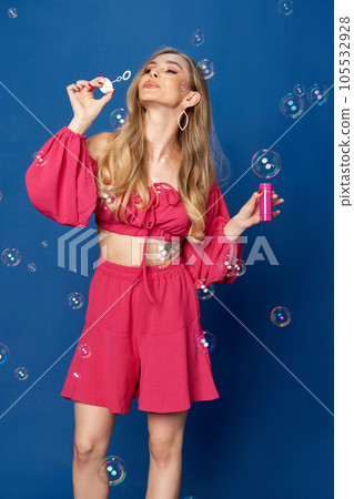 Portrait of blonde girl bright pink suit blowing bubbles and looking away over blue studio background. 105532928