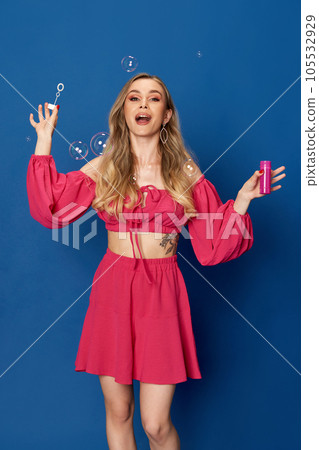 Portrait of excited blonde girl dressed bright pink suit blowing bubbles and looking at camera over blue studio background. Portrait of excited blonde girl dressed bright pink suit blowing bubbles and looking at camera over blue studio background. 105532929