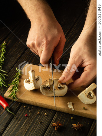 Slicing mushrooms with a knife by the hands of a cook on a cutting board. Cooking mushrooms on the kitchen table with spices and rosemary. 105533949