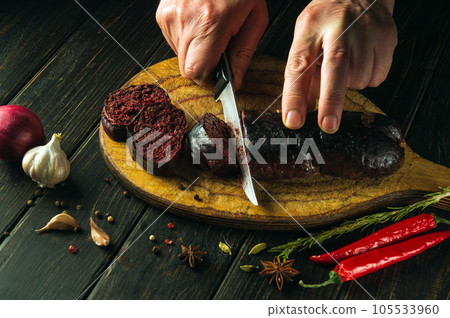 Close-up cook's hands with a knife cut blood sausage on a kitchen cutting board. Cooking a national dish with spices at home in the kitchen 105533960