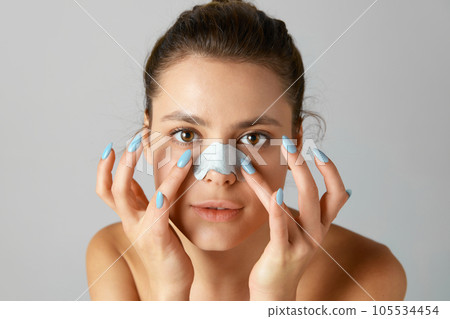 Beautiful young woman using nose mask for cleansing pores against grey studio background. Skin care routine 105534454