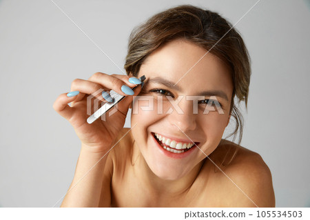 Beautiful young woman taking care after look, appearance plucking eyebrows with tweezer against grey studio background Beautiful young woman taking care after look, appearance plucking eyebrows with tweezer against grey studio background 105534503